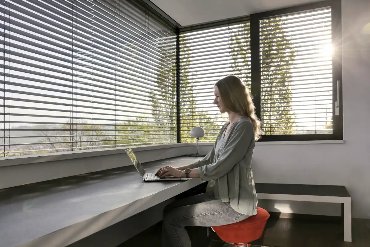 Frau arbeitet an einem Laptop in einem modern eingerichteten Büro mit Jalousien