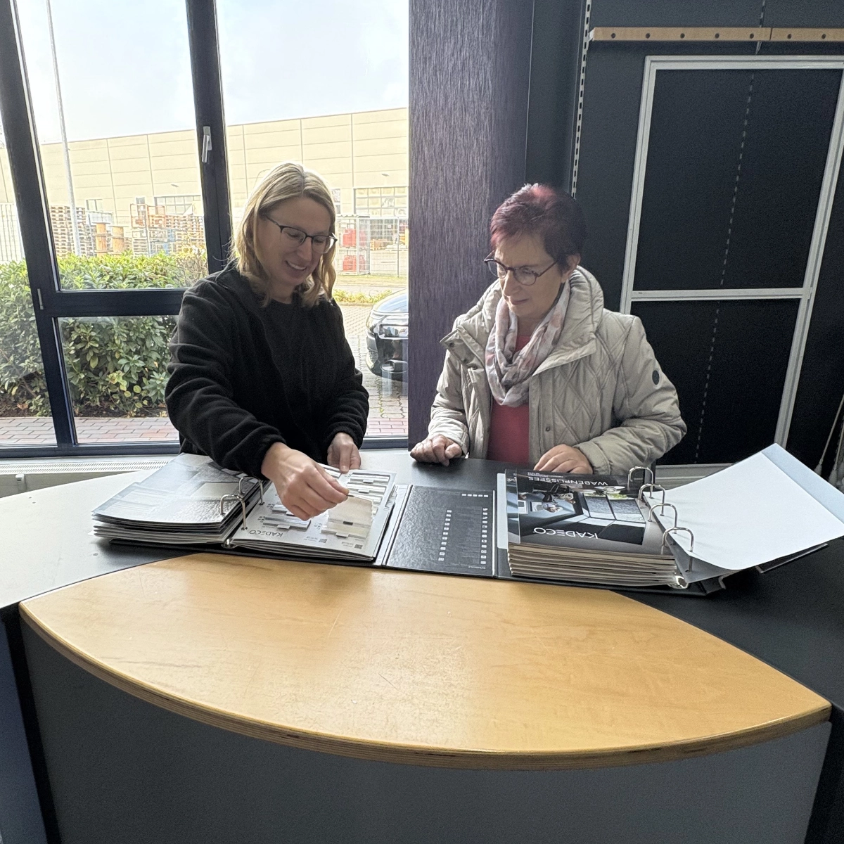 Zwei Frauen an einem großen Tisch. Die eine zeigt der anderen Stoffmuster in einem großen Ordner